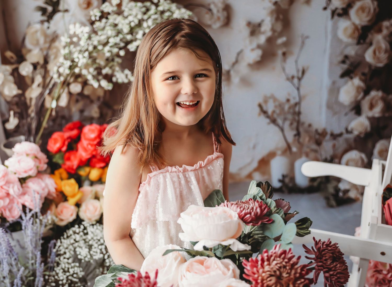 Smiling young girl in pink dress holding white flowers surrounded by colorful floral arrangements in a bright studio setting.