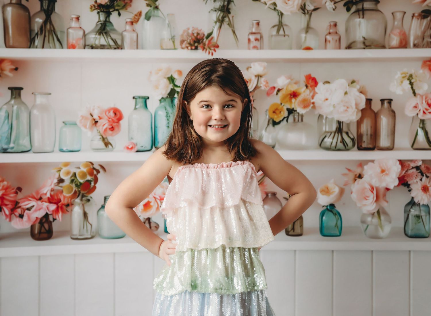 Young girl in ruffled dress poses with hands on hips before white shelves with colorful flowers and glass vases.