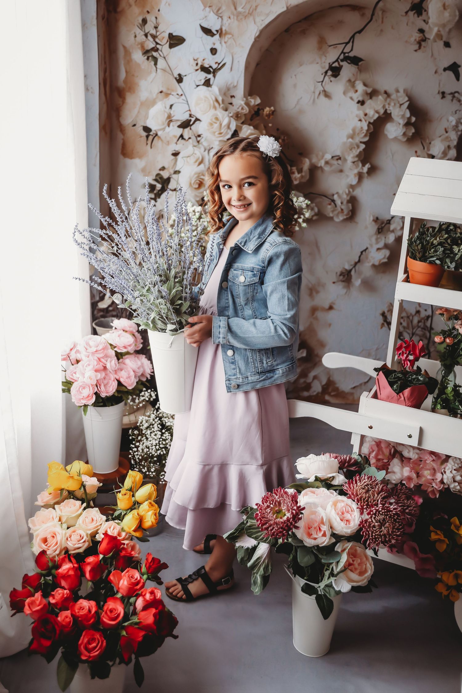 Young girl in denim jacket and pink dress holding lavender bouquet surrounded by colorful flowers in a floral shop setting.