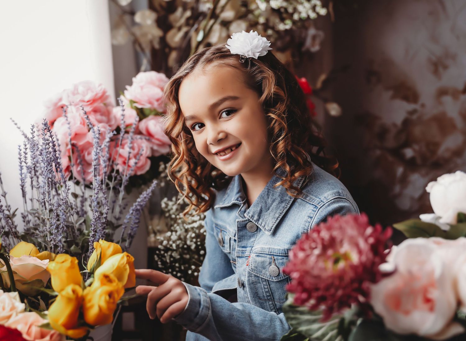 Young girl with curly hair and white flower accessory smiling among colorful blooms in a floral shop setting.