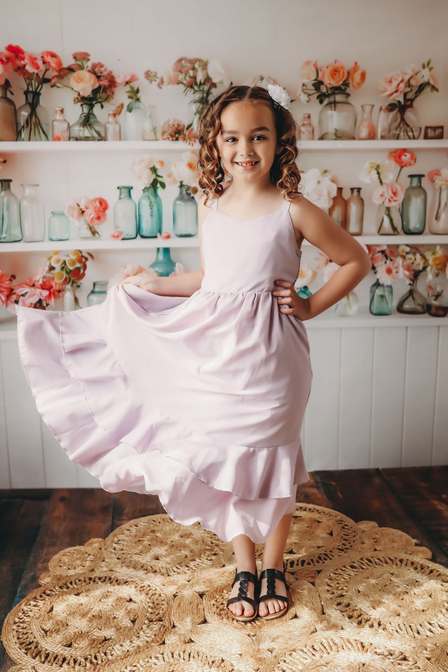 A young girl twirls in a lavender dress with sandals on a woven rug, surrounded by colorful flower vases on white shelves.