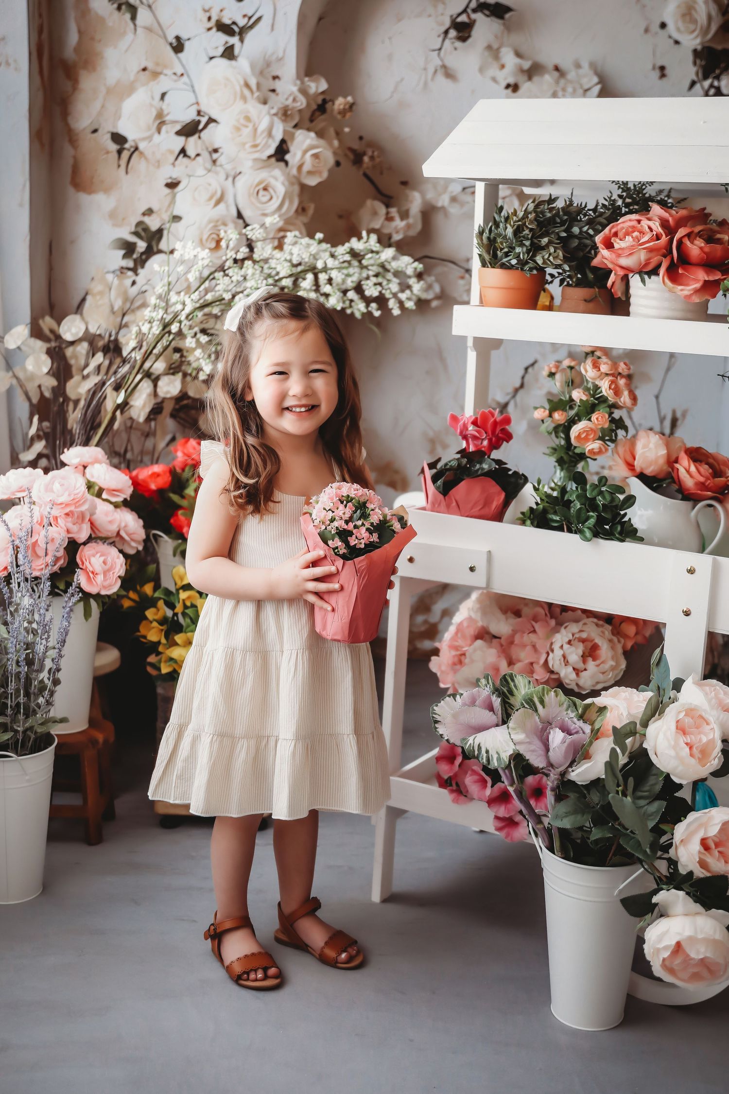 Smiling little girl in white dress holding pink potted flower surrounded by colorful floral arrangements in bright studio.