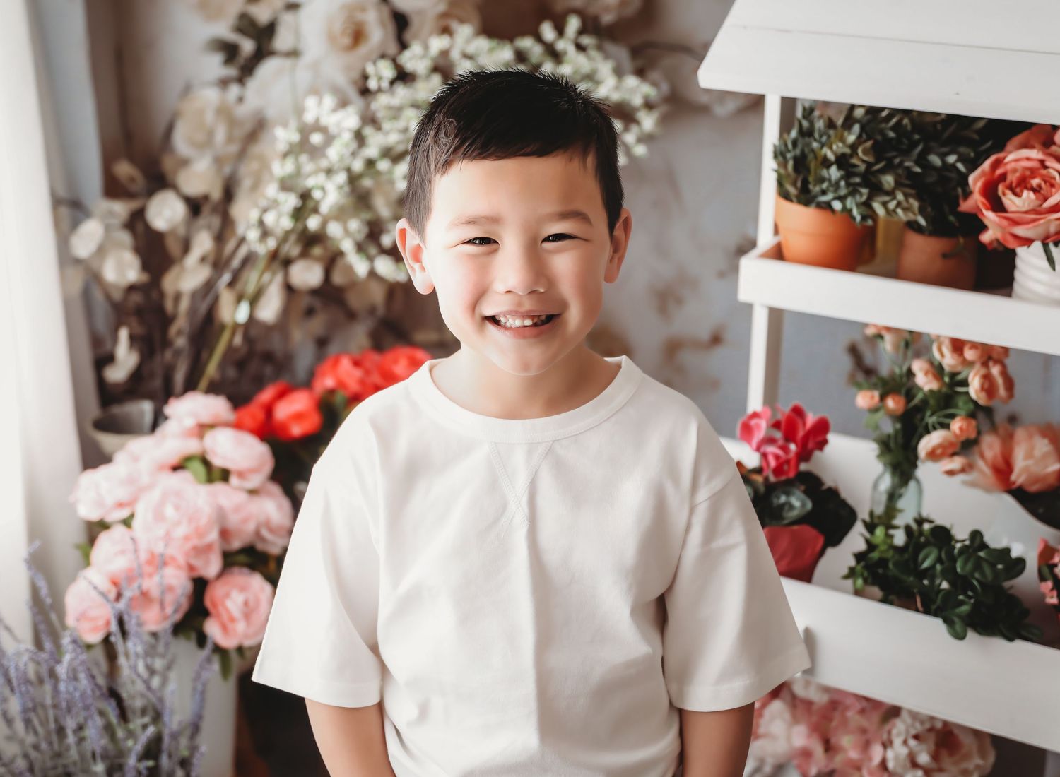 Smiling Asian boy in white shirt poses cheerfully among colorful floral arrangements and potted plants backdrop.