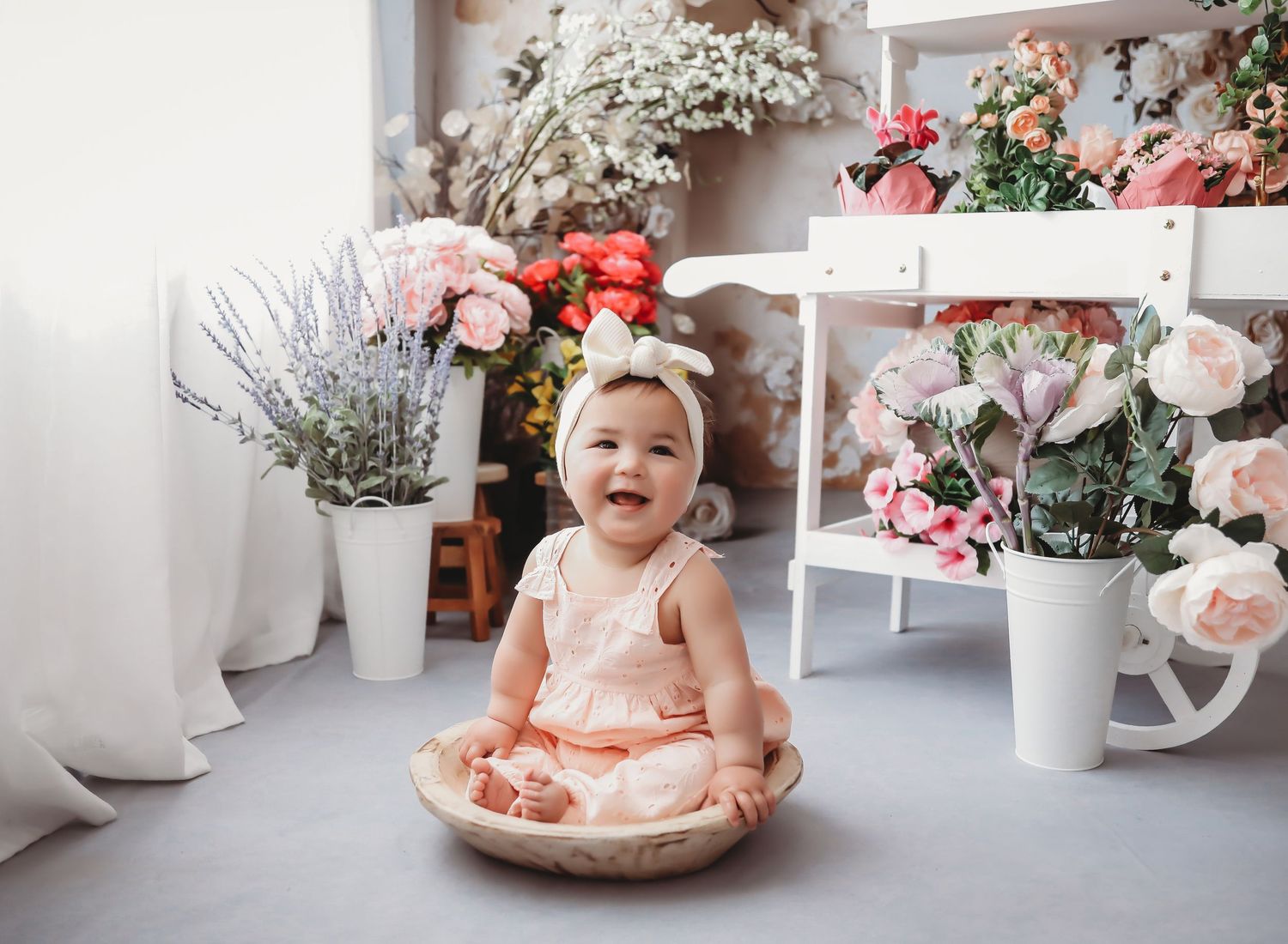 A happy baby girl in a pink dress and bow headband sits in a wooden bowl surrounded by colorful flowers.