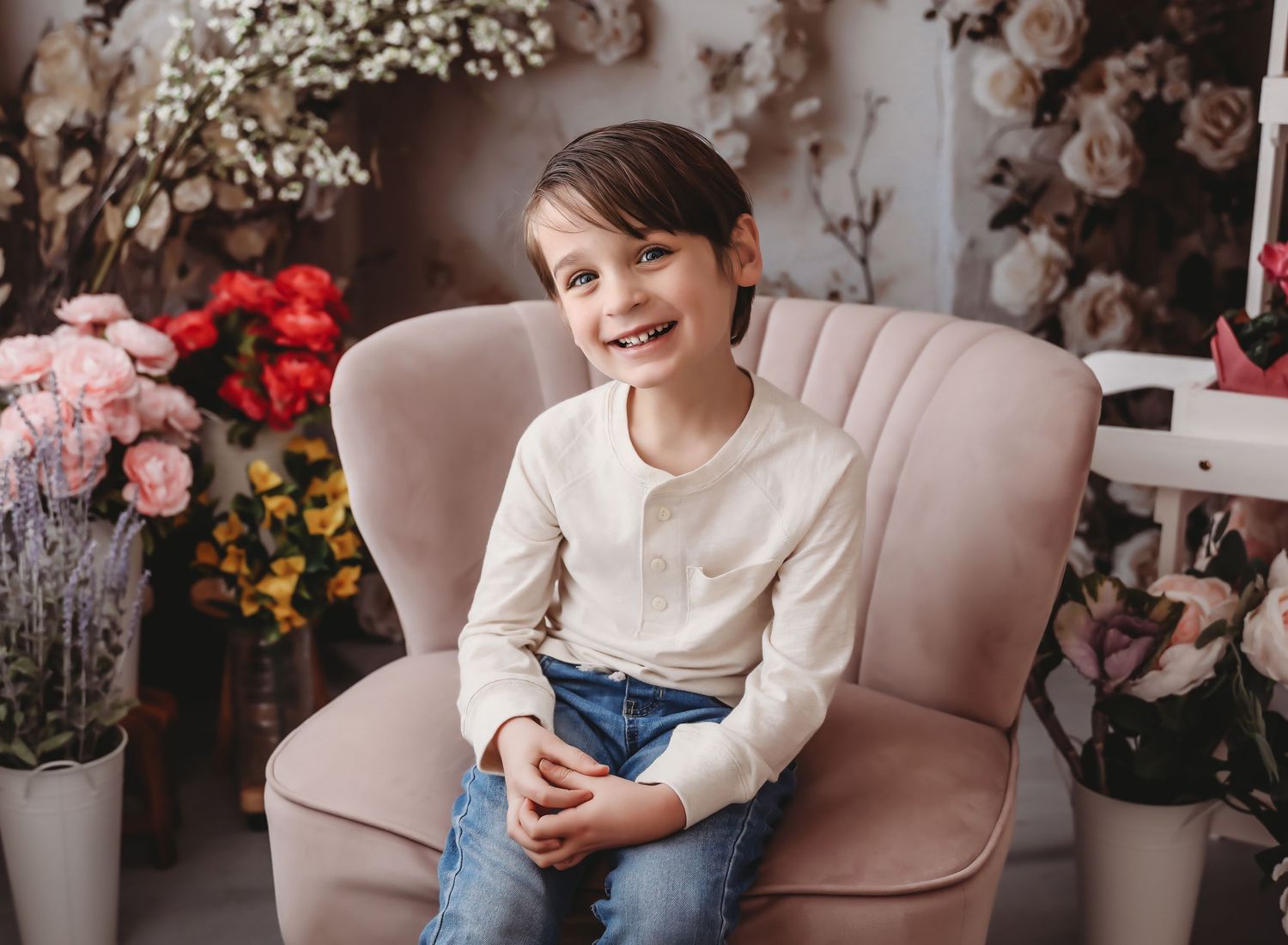 Smiling boy in white shirt and jeans sitting in pink chair surrounded by colorful flowers in studio.