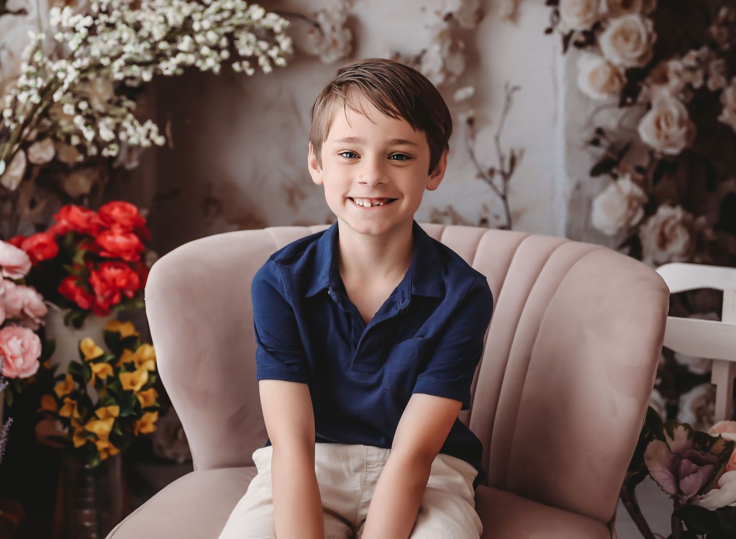 Smiling boy in navy polo shirt sitting in pink chair surrounded by floral backdrop for portrait photo.