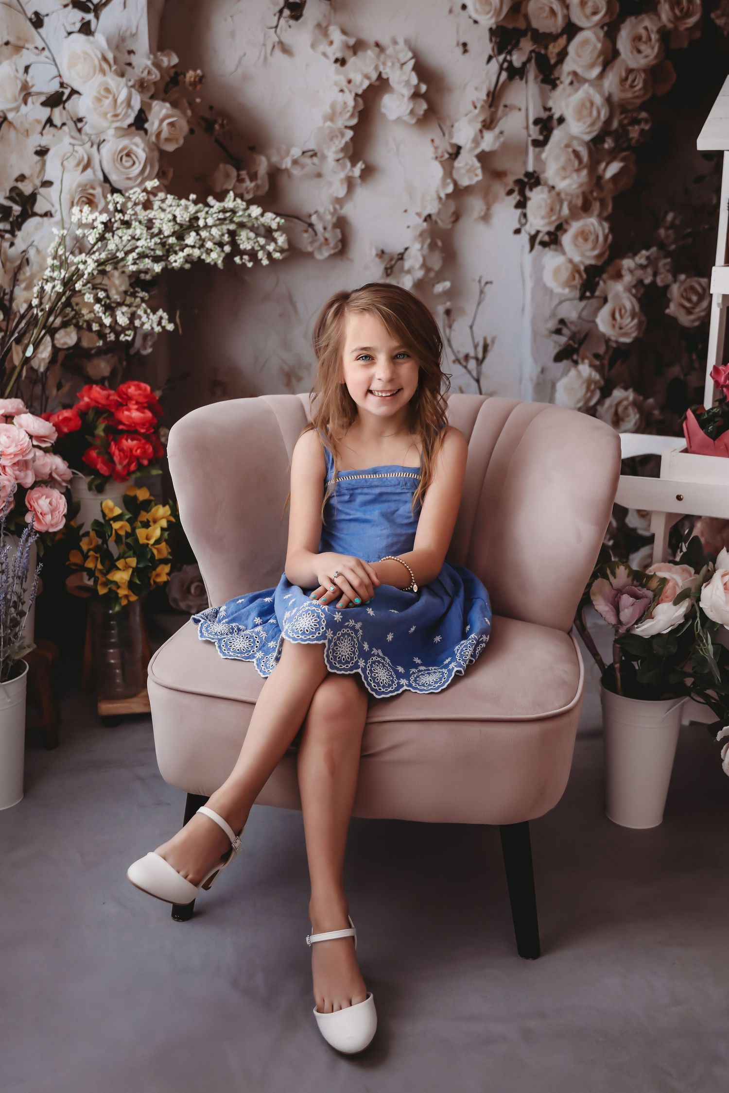 Happy girl in blue dress and white shoes sitting in pink chair with floral studio backdrop.