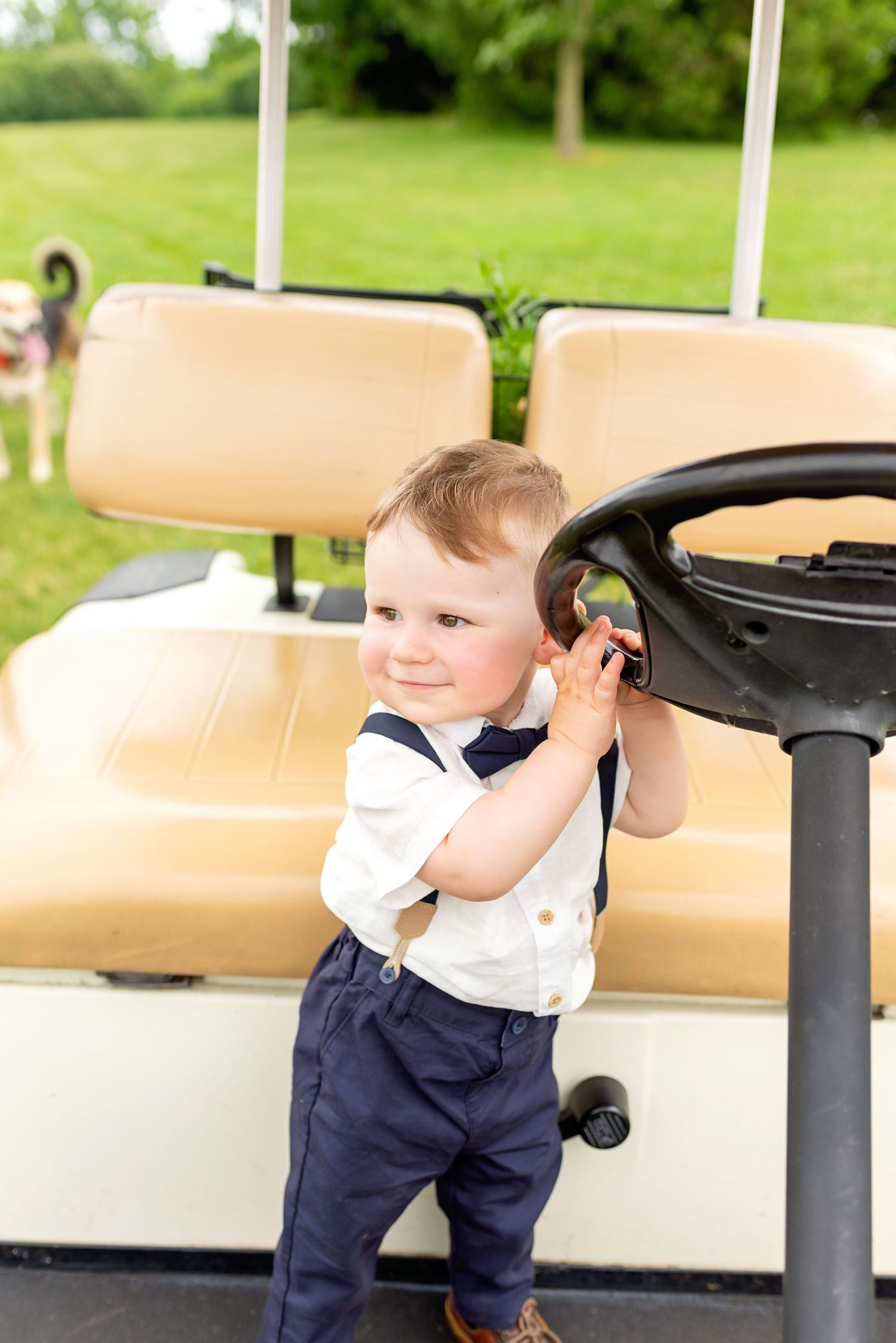 Adorable toddler boy in suspenders playing with golf cart steering wheel outdoors on green grass.