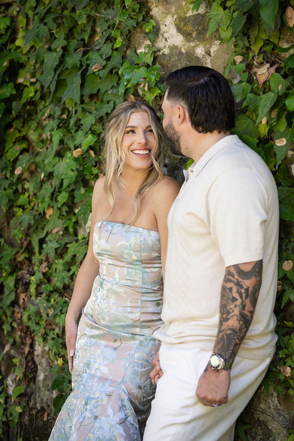 Smiling blonde woman in floral strapless dress with tattooed man in cream outfit against ivy-covered stone wall.