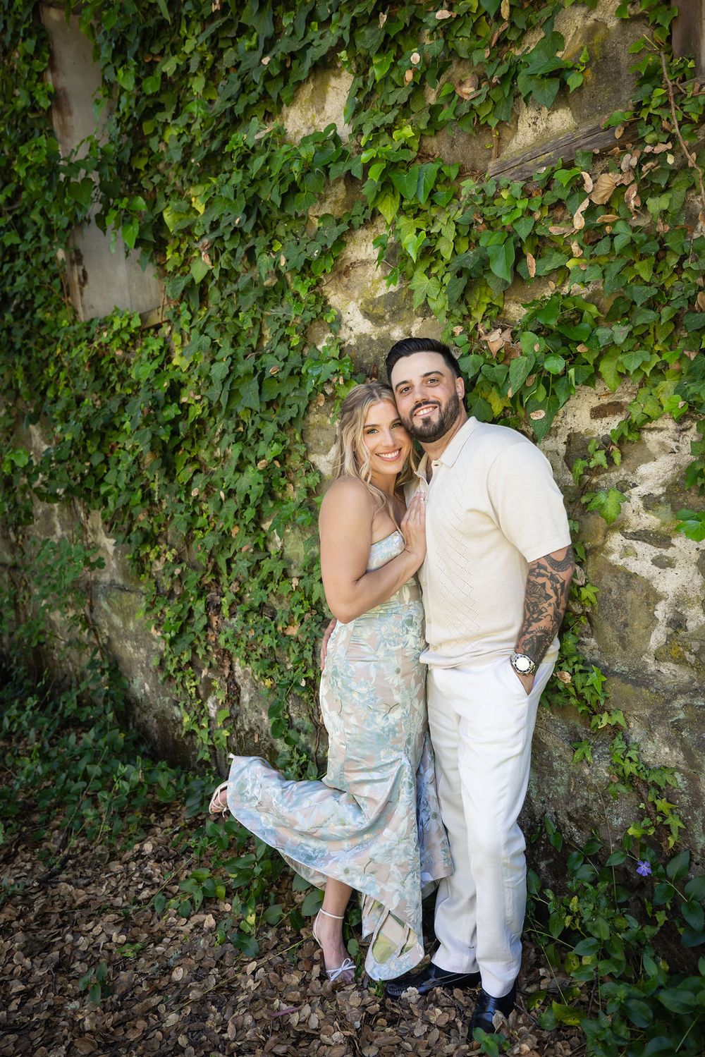 Happy couple posing together in front of lush ivy stone wall, woman in floral dress and man in white outfit.