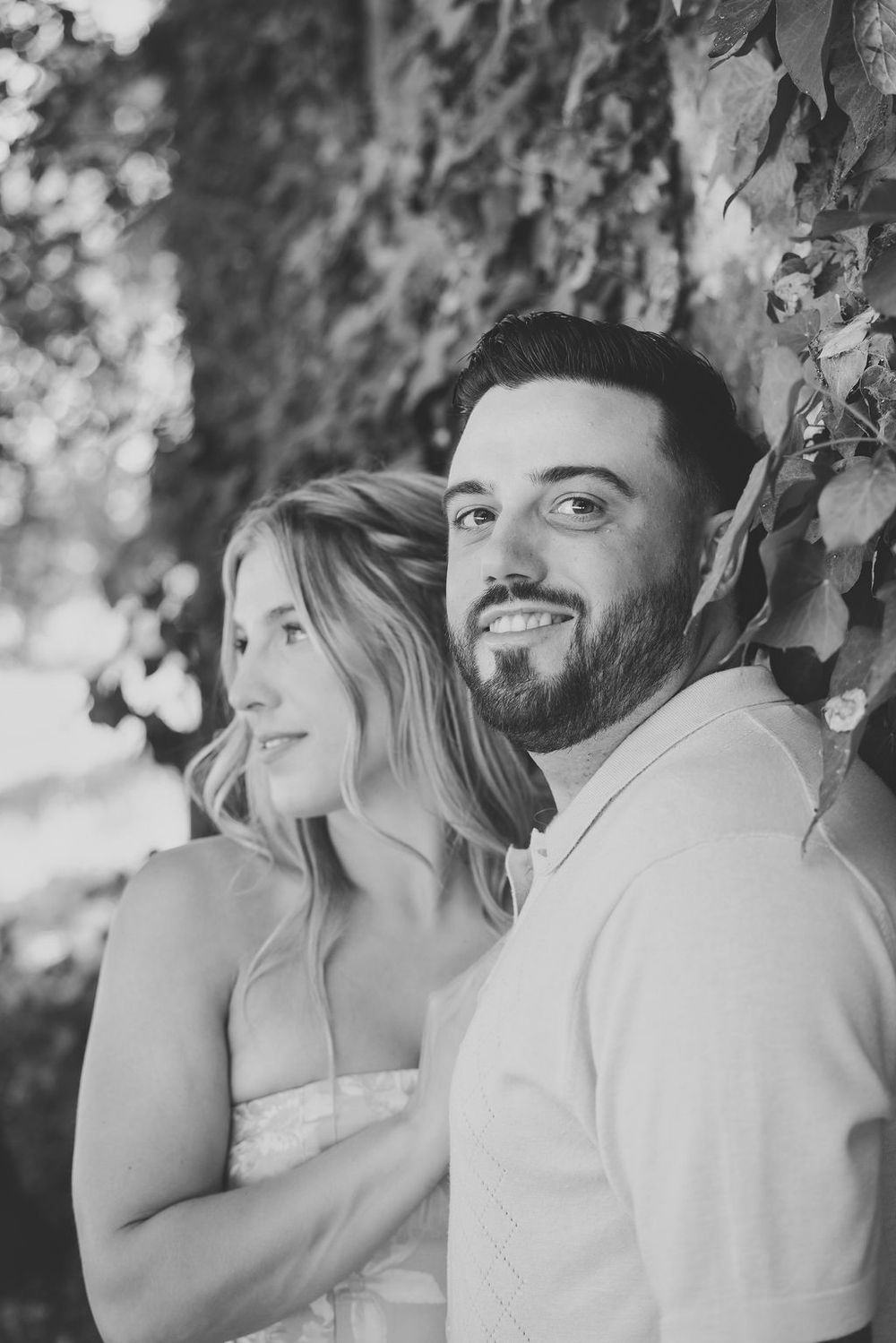 Black and white portrait of a smiling bearded man and blonde woman posing together under a leafy tree.