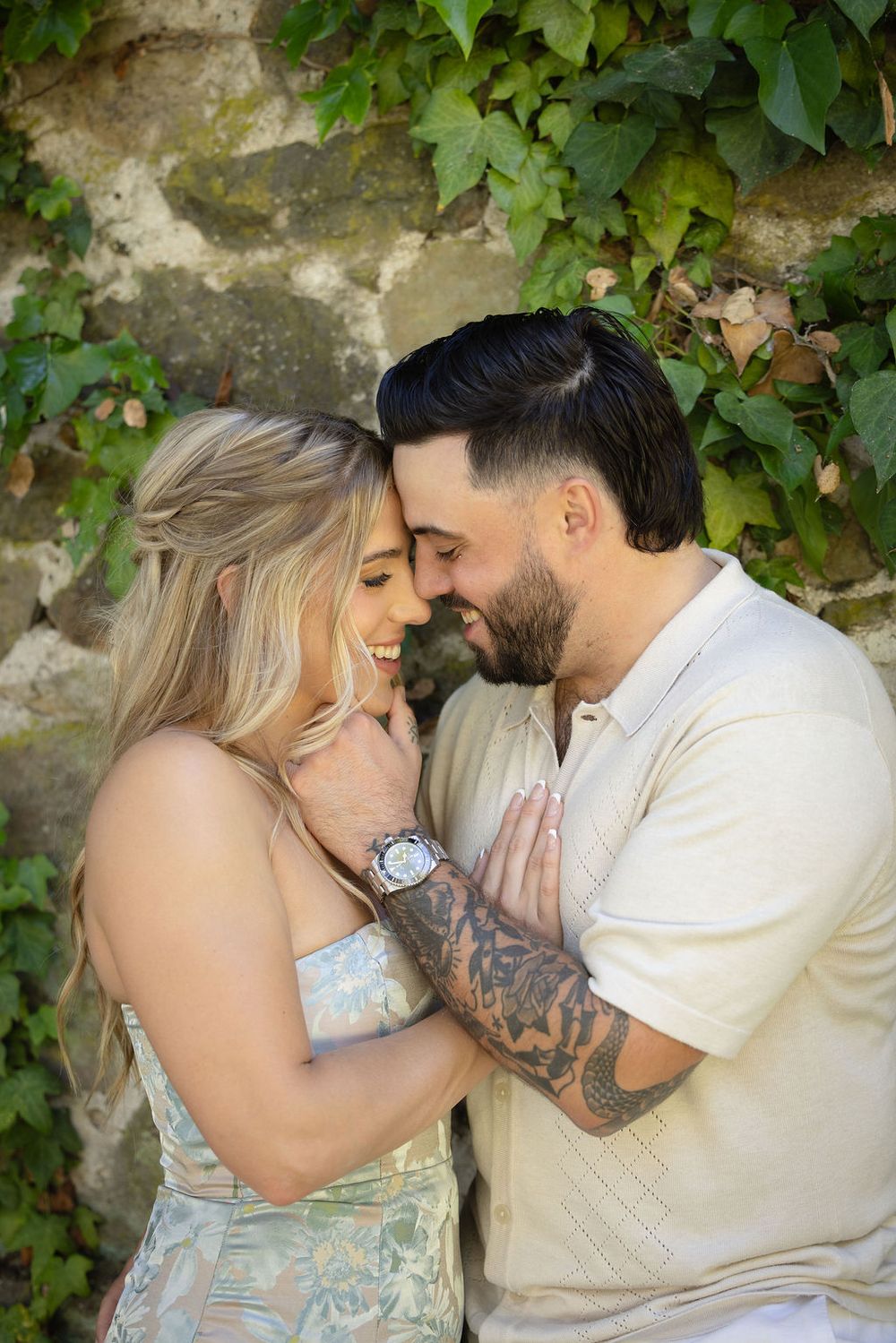Romantic couple sharing an intimate moment, foreheads touching and smiling, against a stone wall covered in ivy.