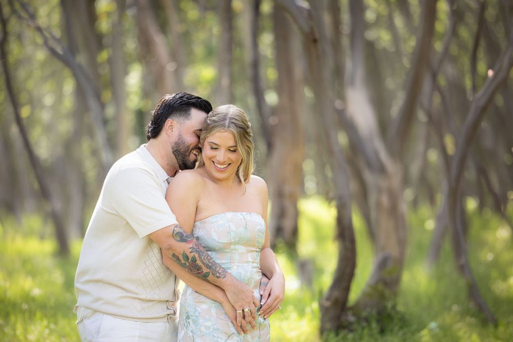 Couple sharing a tender moment in a sunlit forest, man with tattoos embracing pregnant woman in a floral dress.