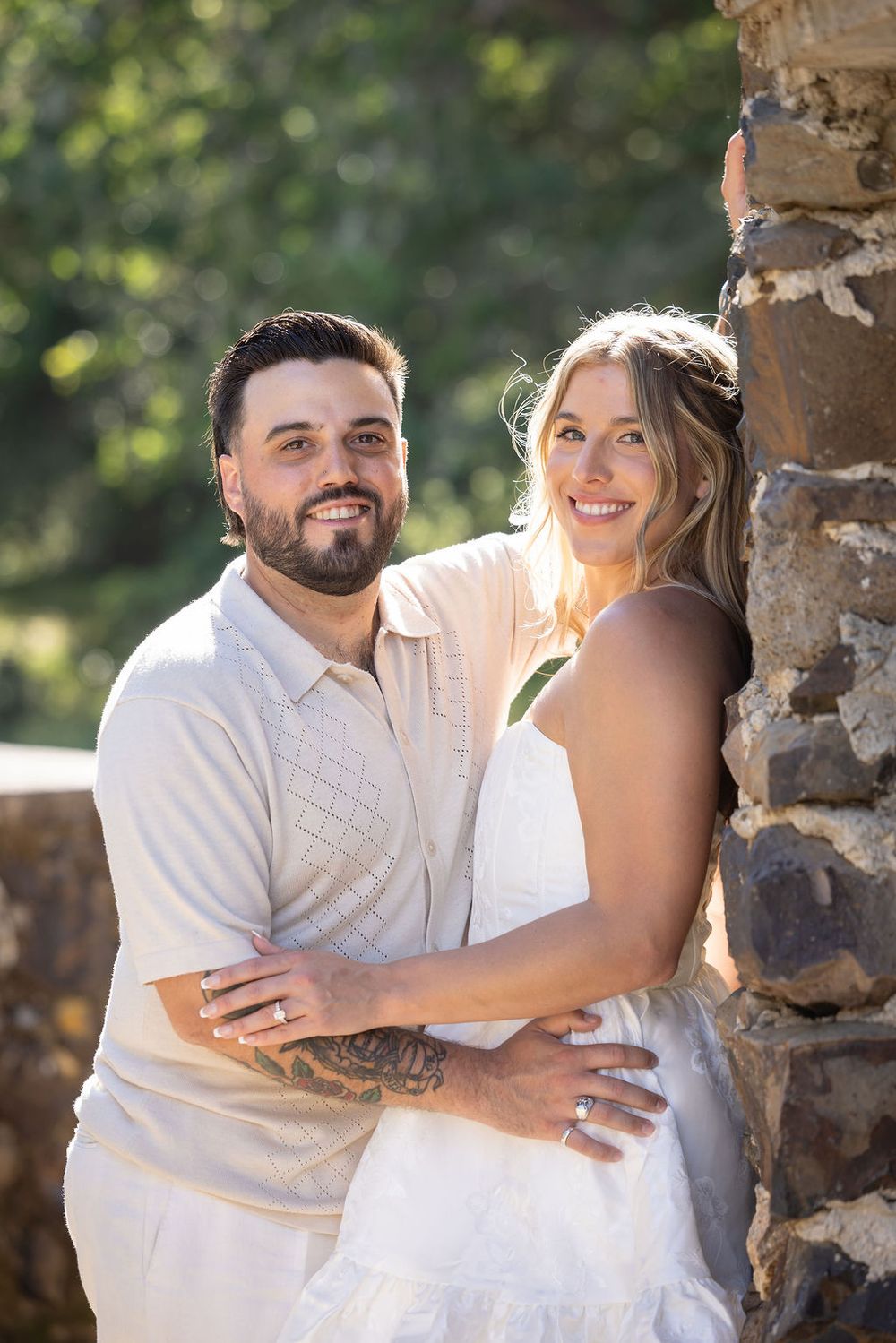 Smiling couple posing by rustic stone wall outdoors, man with tattoos embracing woman in white dress.