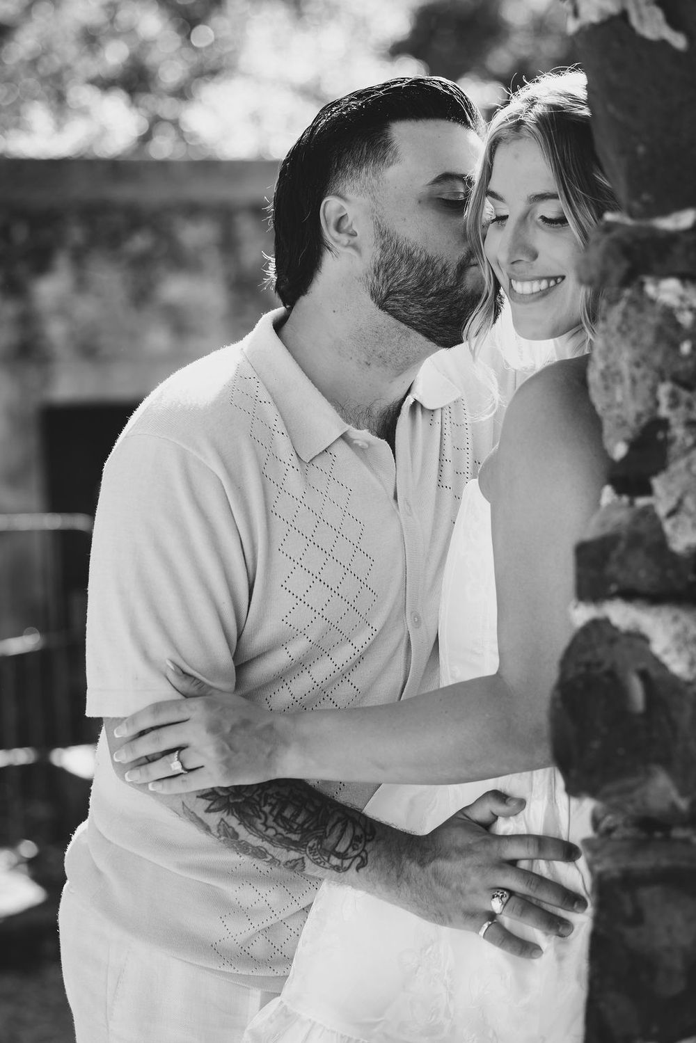 Black and white photo of a man with tattoos kissing a smiling woman on the cheek near a stone wall.