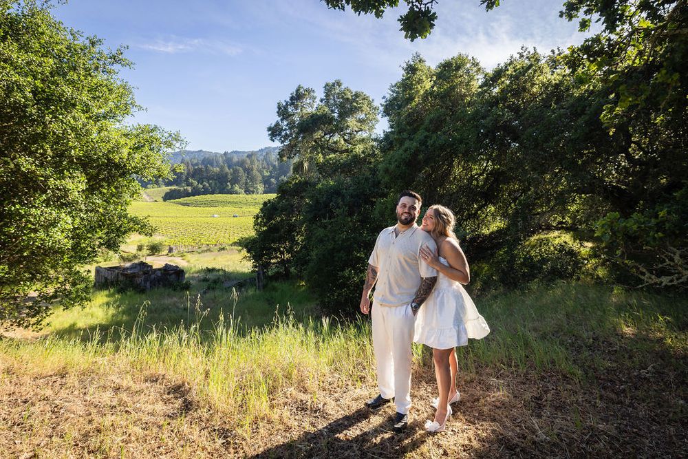 Happy couple in white outfits standing together on a sunny hillside surrounded by lush green trees.