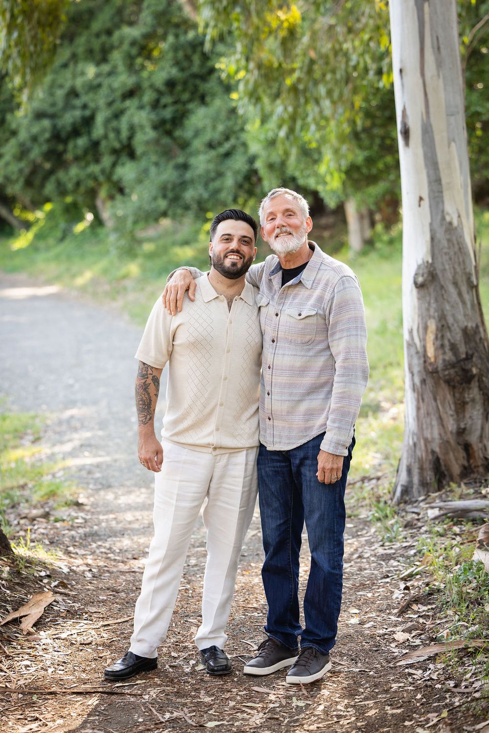 Two men smiling and posing together on a tree-lined path, one with tattoos in beige outfit.