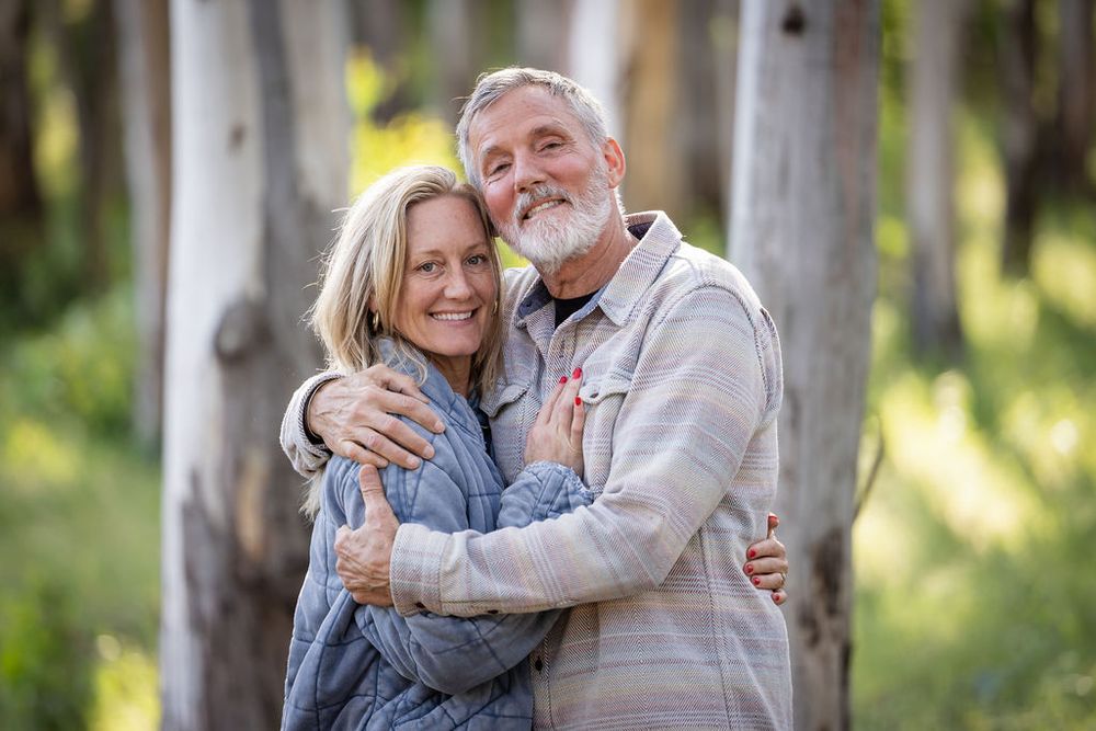 Older couple embracing warmly among tall trees in a sunlit forest setting.