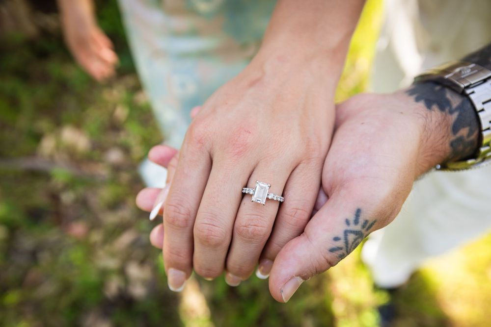 Close-up of two hands holding, one wearing a sparkling emerald-cut diamond engagement ring, with a tattooed wrist visible.