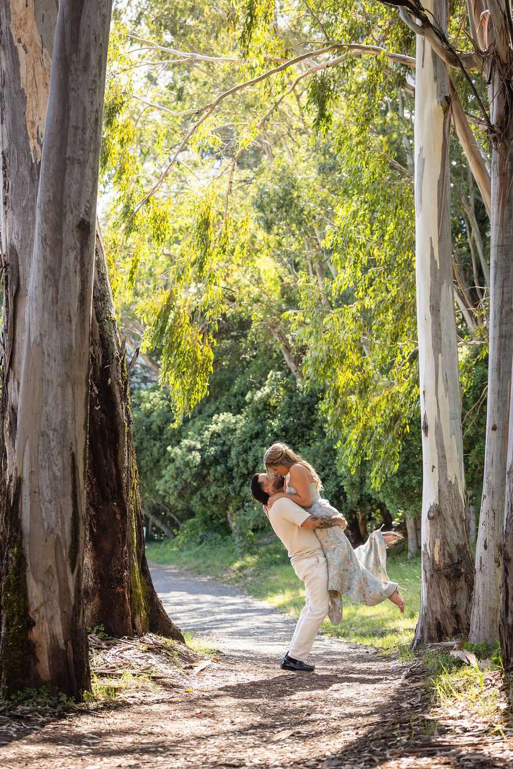 Romantic couple sharing a lift and kiss on a sunlit tree-lined path surrounded by lush green eucalyptus trees.