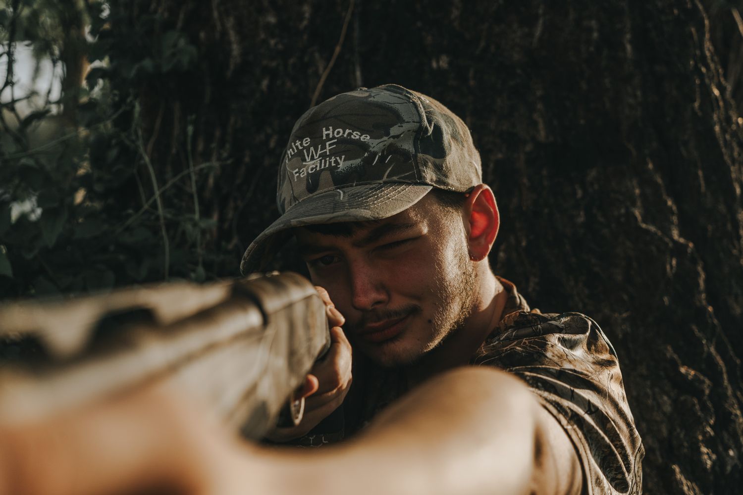 Man in camo cap aiming a rifle outdoors near a tree in a hunting or tactical setting.