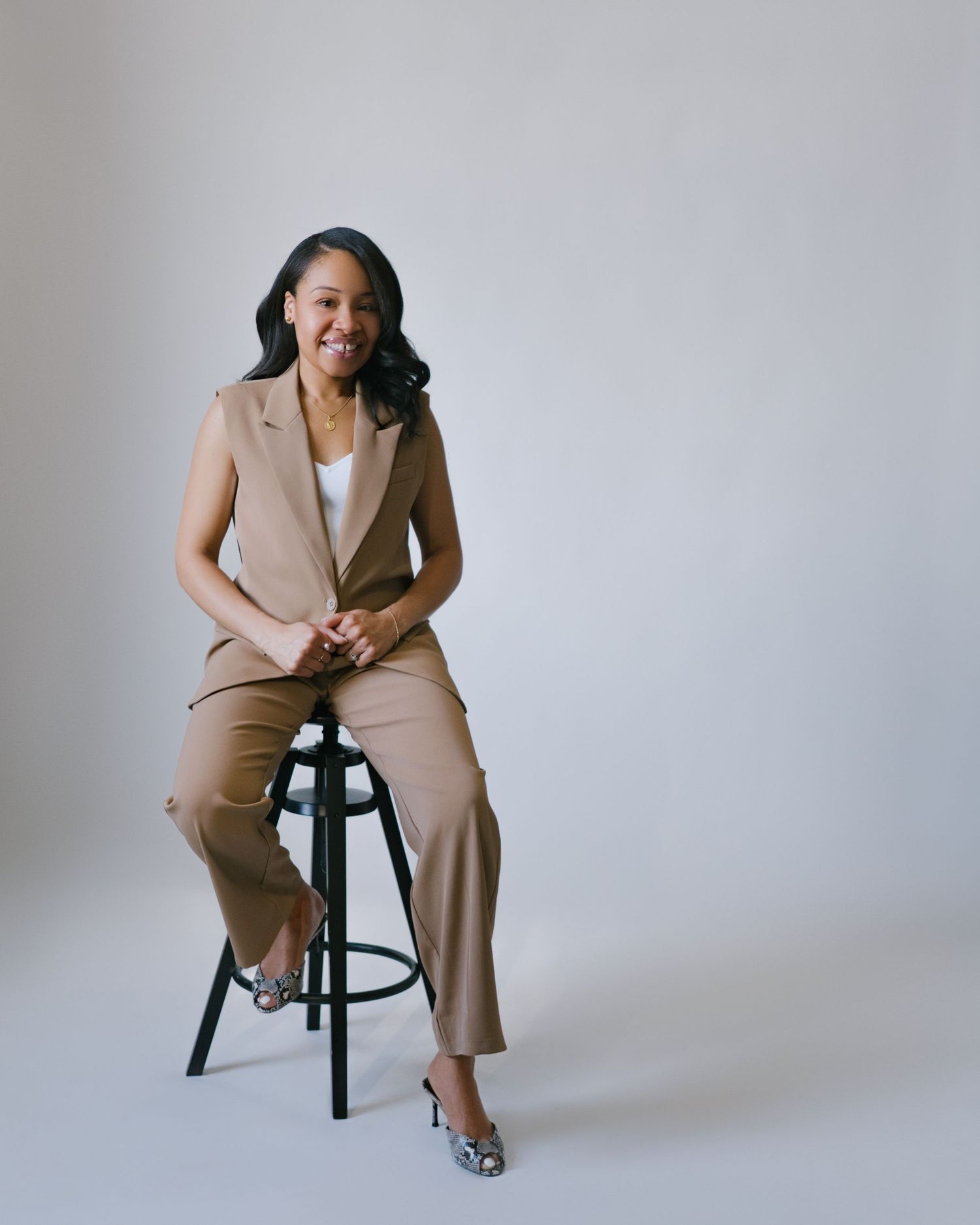 Woman in tan sleeveless suit sitting on stool, smiling confidently in a bright studio setting.
