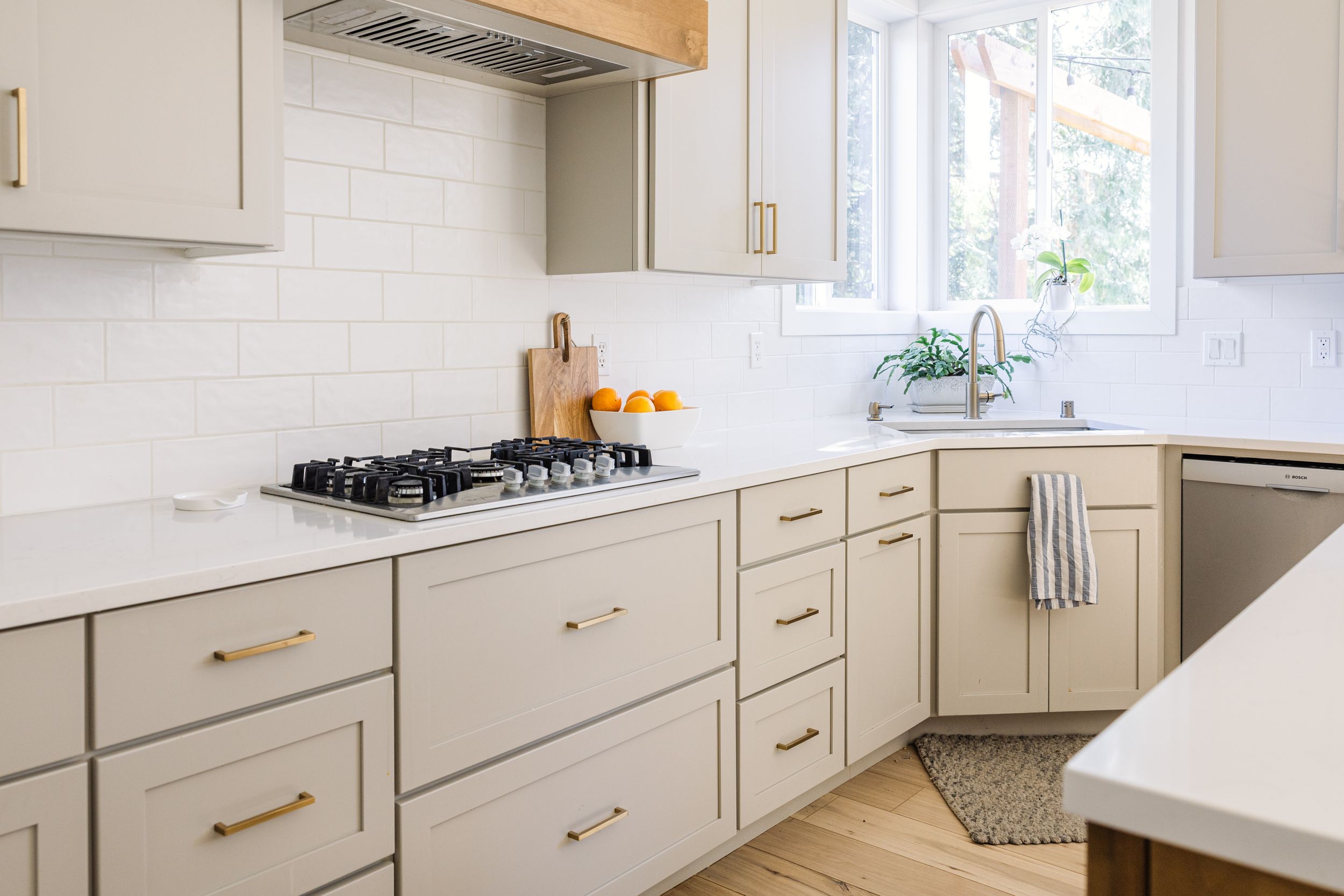 Modern kitchen with light gray shaker cabinets, brass hardware, white subway tile backsplash, and stainless steel gas cooktop.