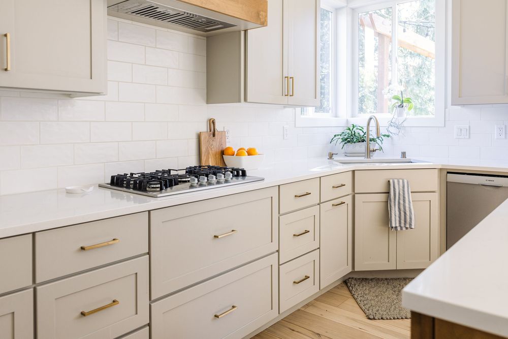 Modern kitchen with light gray shaker cabinets, brass hardware, white subway tile backsplash, and stainless steel gas cooktop.