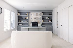Elegant home office with gray built-in shelving, framed gallery wall, curved desk, and white sofa seating area.