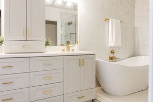 Luxurious white bathroom with gold hardware, freestanding soaking tub, marble tile walls, and gray vanity cabinets.