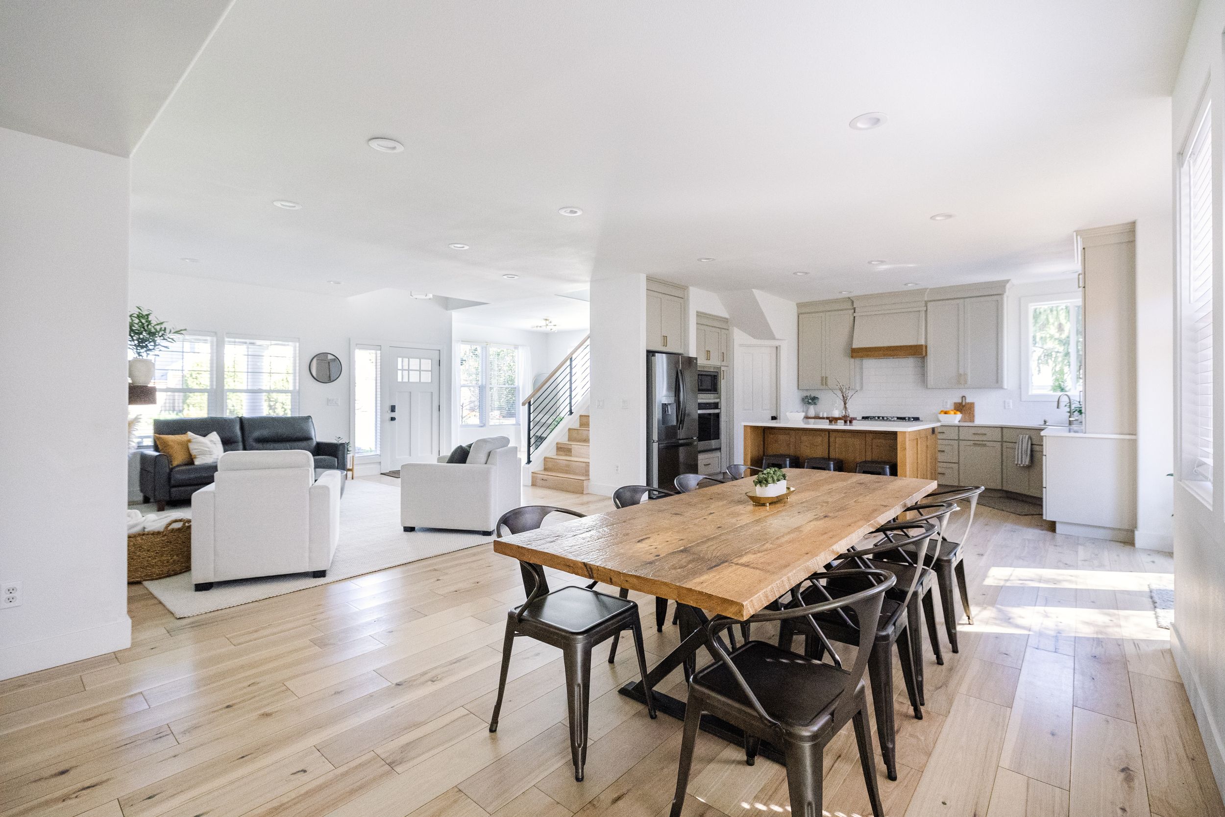 Open-concept dining and living area with rustic wood table, light hardwood floors, and modern white kitchen beyond.