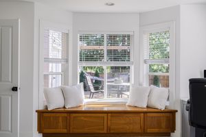 Bright bay window seating area with white cushions, wooden storage bench, and plantation shutters.
