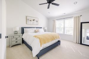 Bright master bedroom with navy upholstered bed, ceiling fan, large window, and neutral carpet flooring.