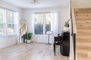 Bright entryway with hardwood floors, black piano, staircase, and large windows with white curtains.