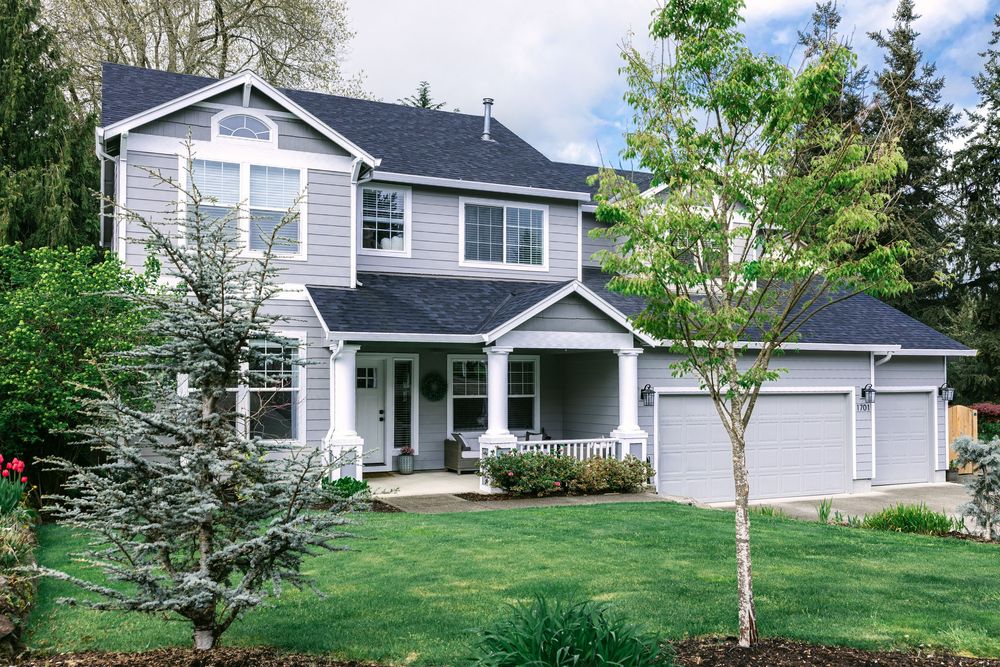 Two-story gray craftsman home with covered porch, attached garage, and lush green landscaping.