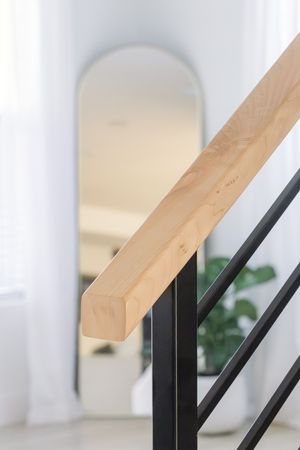 Modern staircase with light wood handrail and black metal balusters, with an arched mirror visible in background.