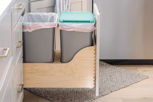 Pull-out double trash can drawer with gray bins in kitchen cabinet next to stainless steel refrigerator.