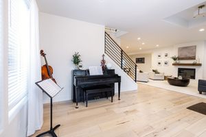 Bright music room with black upright piano, violin, and music stand beside modern staircase and fireplace.