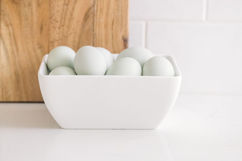 White ceramic bowl filled with pale green eggs on white kitchen counter with wood cutting board behind.