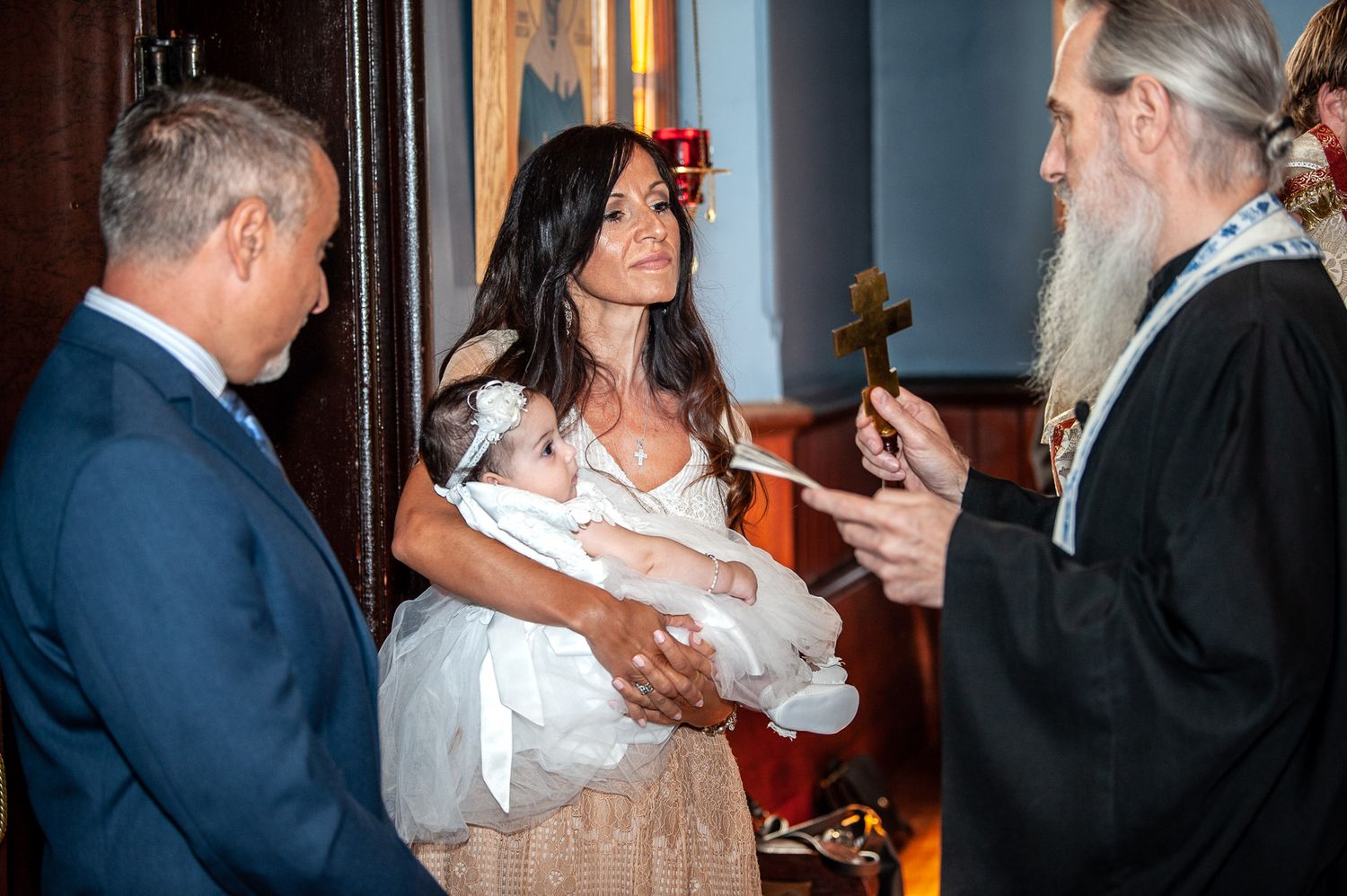 Orthodox priest blessing a baby held by its mother while the father stands nearby in a church.