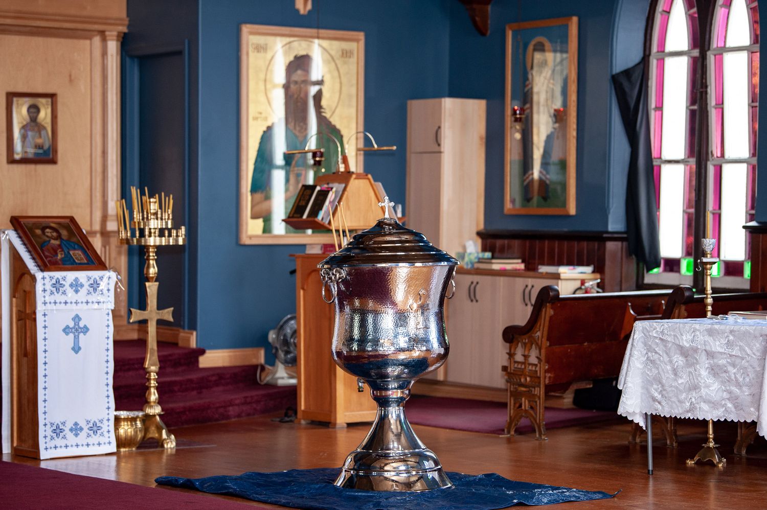 Orthodox church interior with golden baptismal font, iconostasis, and religious icons on blue walls.