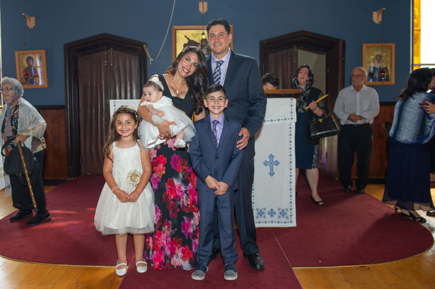 Family portrait at Orthodox church baptism with parents, baby, and two children smiling.