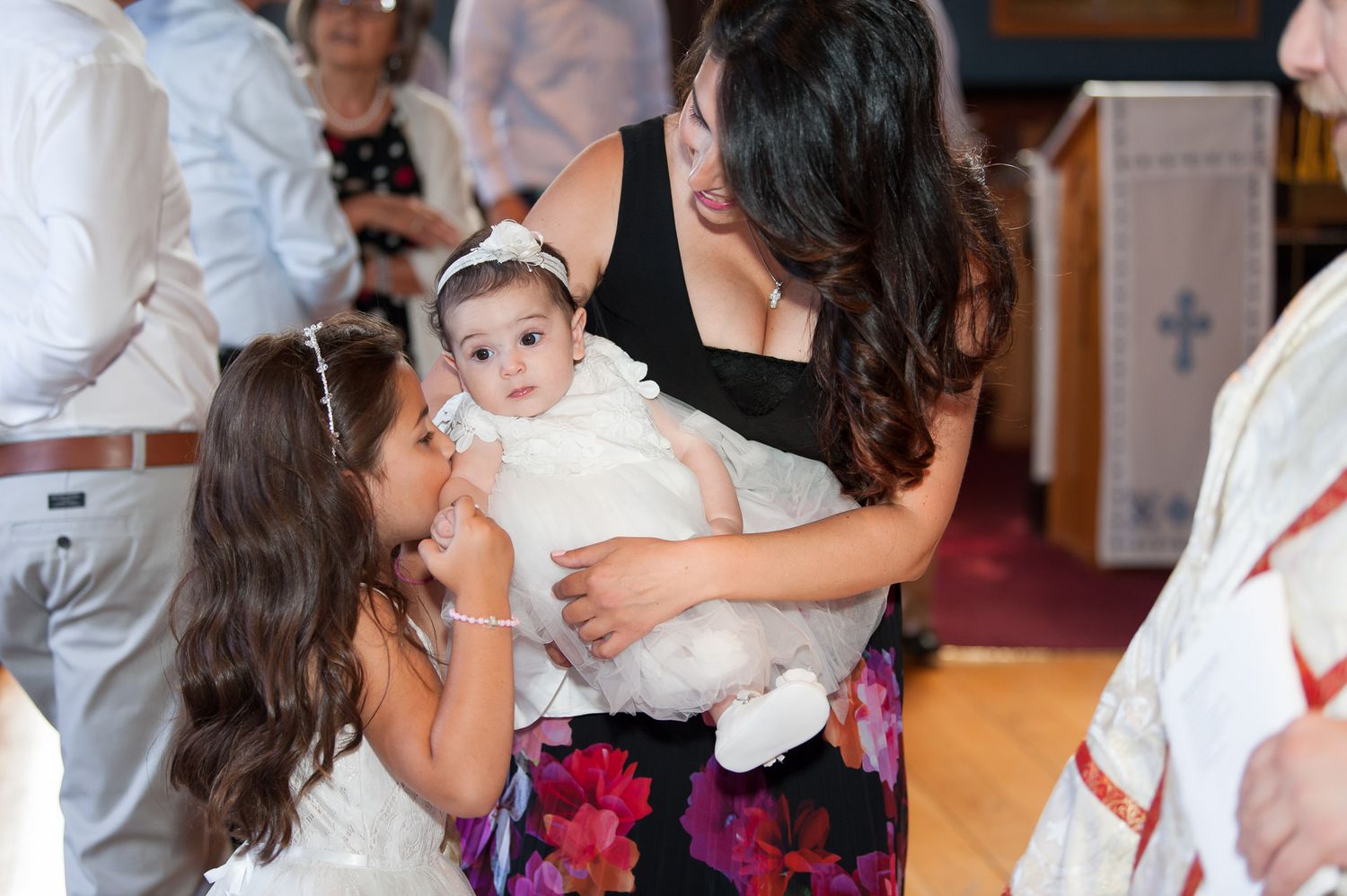 Mother holding baby in white baptism gown while young girl looks on in church.