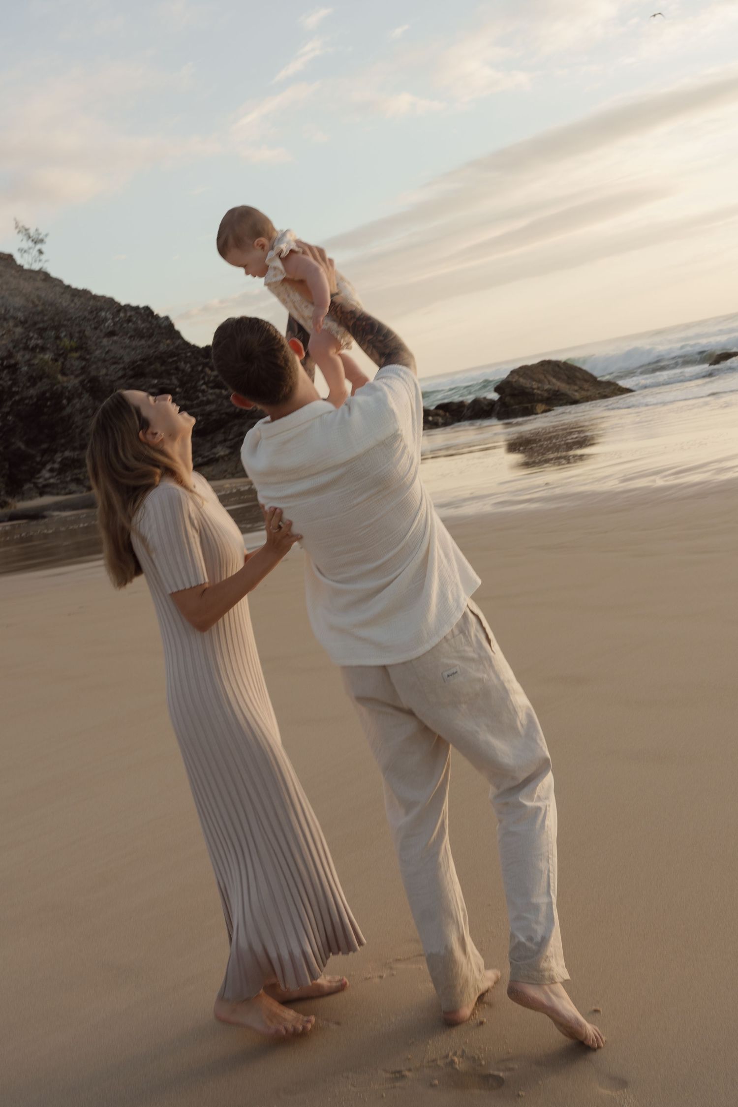 A couple in white outfits lifts their baby joyfully on a sandy beach at sunset.