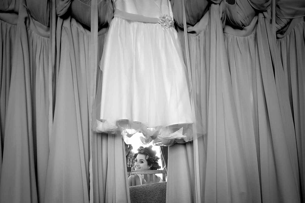 Black and white photo of a wedding dress hanging between draped curtains with a bride peeking through below.