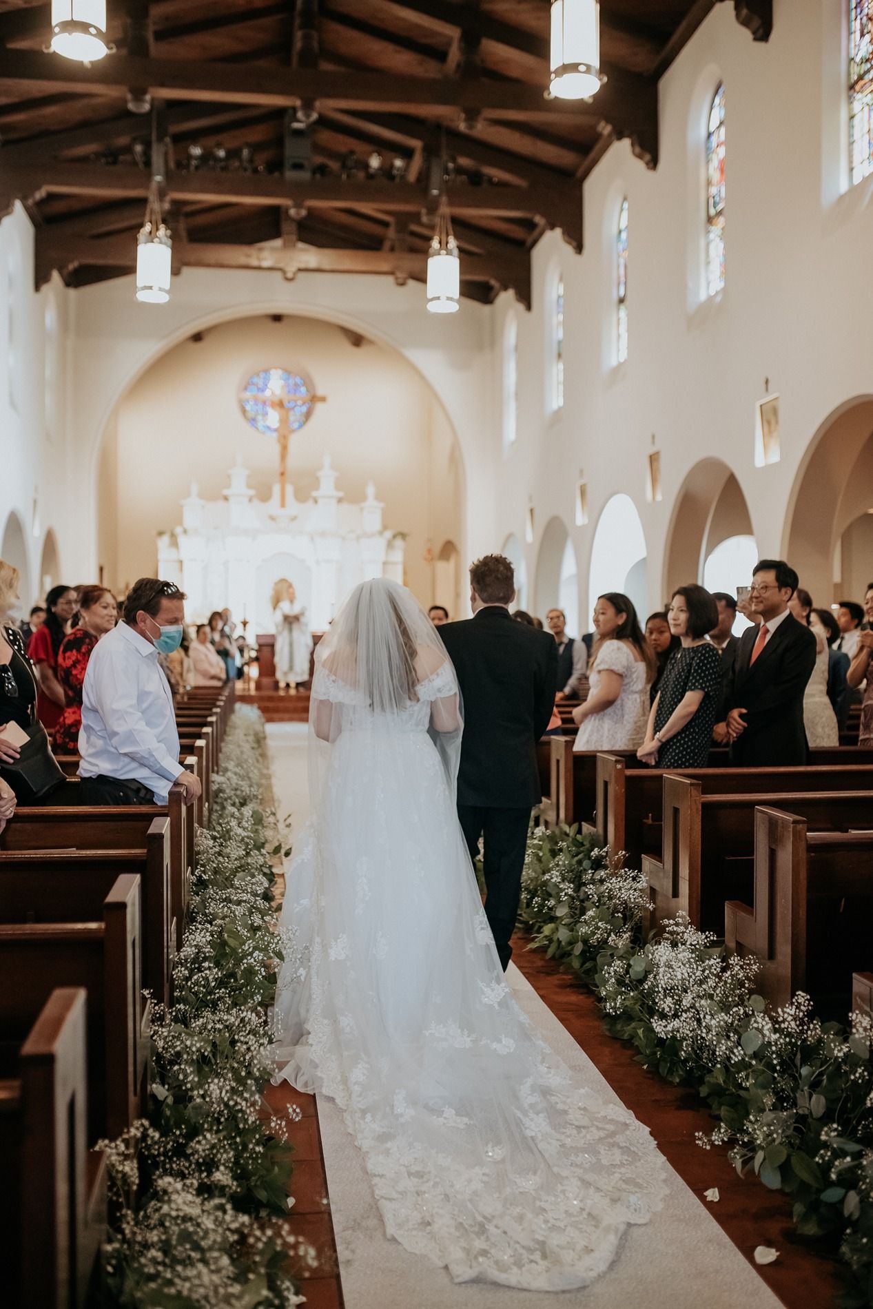 Bride walking down the aisle in a church wedding ceremony, wearing a long veil and gown, flanked by floral arrangements.