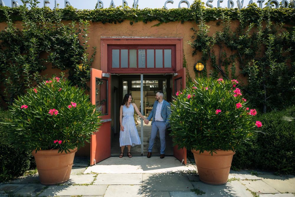 Couple holding hands outside charming red-framed entrance with lush greenery and pink flowering potted plants at Garfield Park conservatory