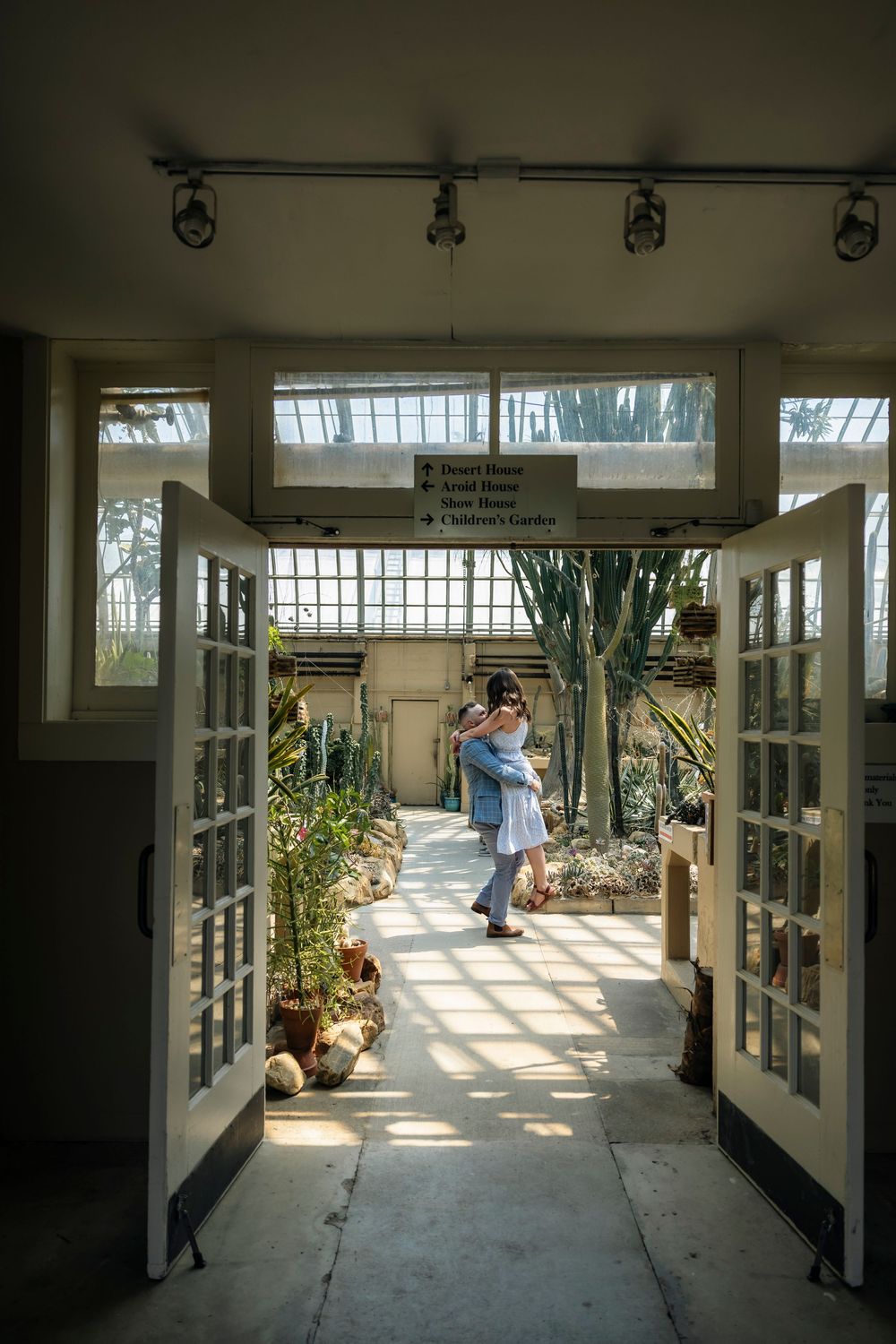 Couple embracing in sunlit greenhouse doorway surrounded by tropical plants and cacti in Chicago Garfield Park Conservatory