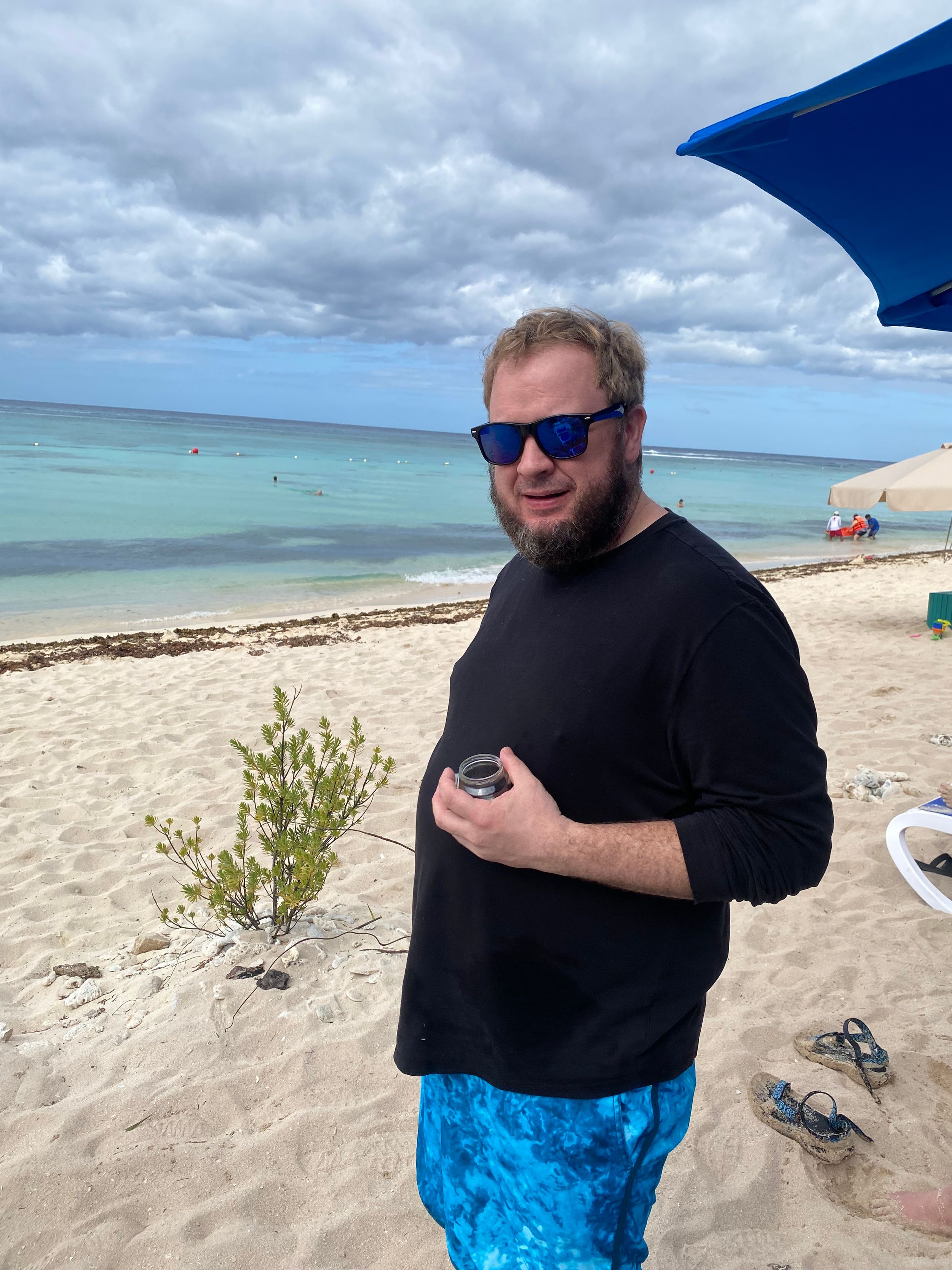 Man in black long-sleeve shirt and blue swim trunks standing on a sandy beach holding a drink on a cloudy day.