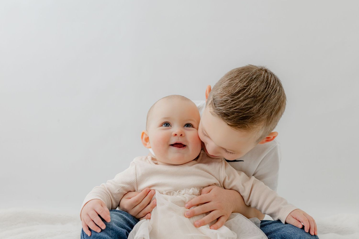 Older boy kissing baby sibling on cheek against white background, sweet family portrait moment.